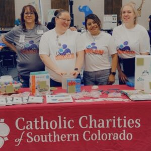 Four staff members from Catholic Charities of Southern Colorado stand behind a display table promoting their Lactation Empowerment Program. The table is covered with educational materials and breastfeeding supplies, with a red tablecloth featuring the organization's name and logo.