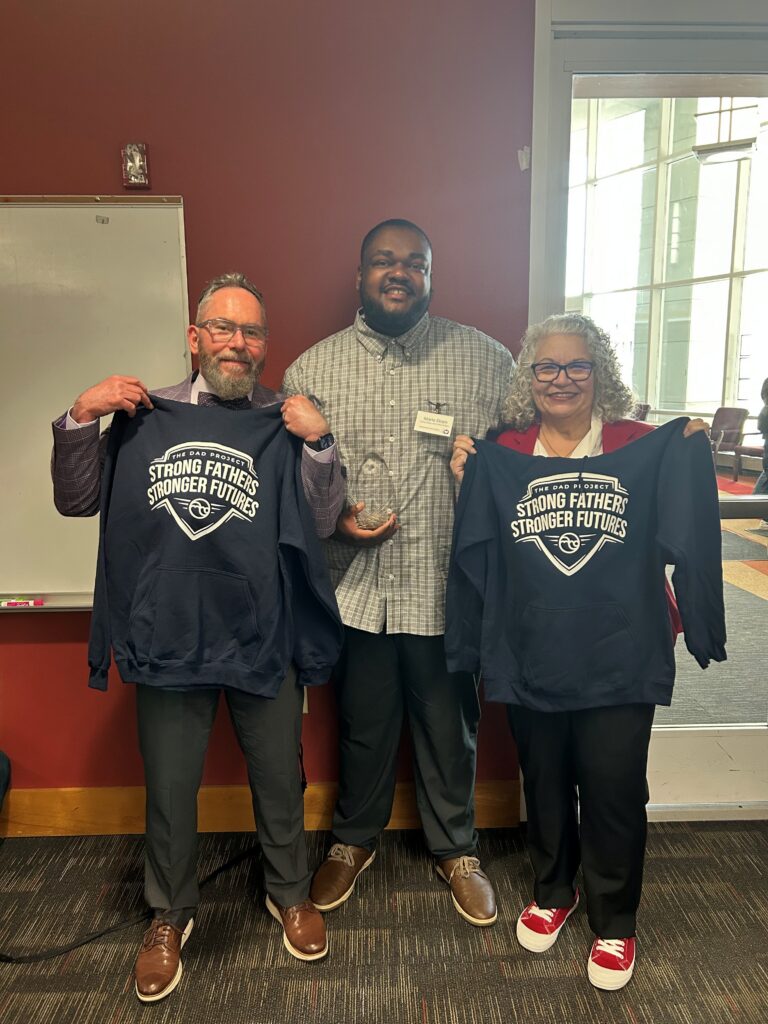 Three smiling people posing indoors; two hold navy hoodies reading "Strong Fathers Stronger Futures" and the center person holds an award.