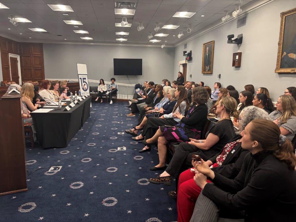 Panel discussion in a conference room with a long table of speakers facing a seated audience, portraits on the walls.