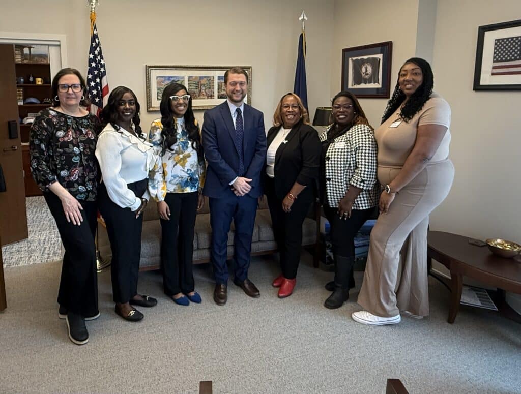 Seven people in business attire standing in an office, smiling in front of flags and framed artwork.