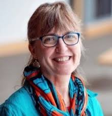 Smiling middle-aged woman with glasses, wearing a teal top and colorful orange-and-blue scarf, in an indoor setting.