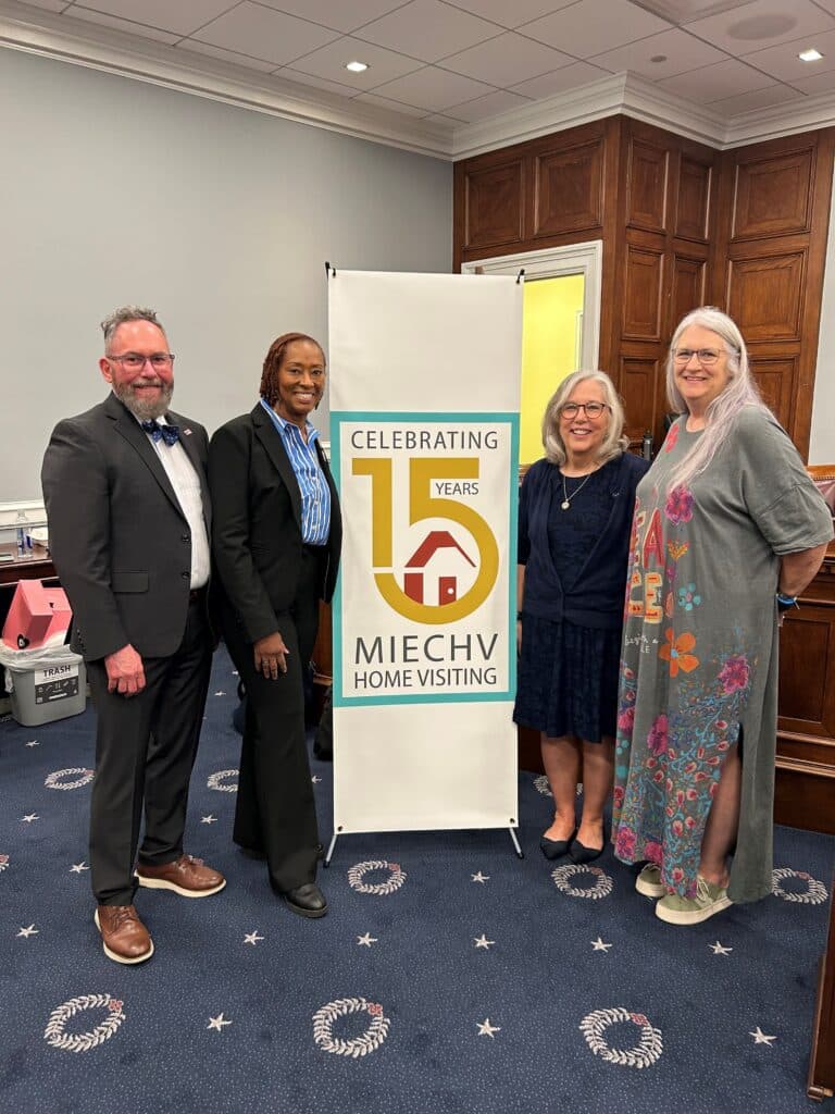 Four smiling adults standing around a banner that reads "Celebrating 15 Years MIECHV Home Visiting" in a meeting room