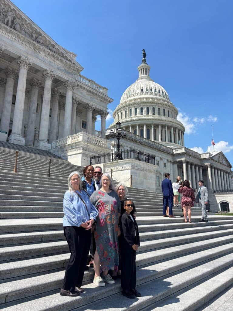 Group of smiling people posing on the Capitol steps with the Capitol dome and classical columns in the background.
