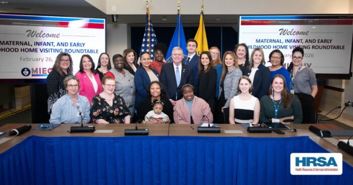 Group of diverse professionals and parents, including a mother holding an infant, posed behind a conference table with flags and screens reading "Maternal, Infant, and Early Childhood Home Visiting Roundtable."