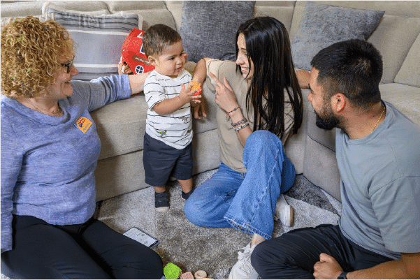 Autism-Within-Families Toddler standing on carpet handing a toy to a smiling woman while three adults sit around them on a living room floor couch, toys scattered.