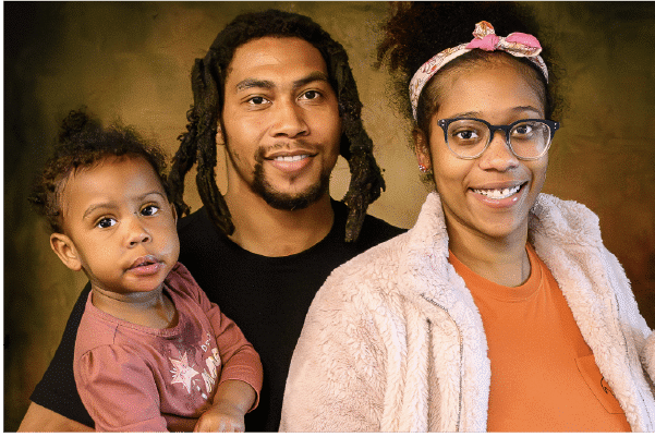 Engaged Smiling family portrait of a man with dreadlocks, a woman wearing glasses and a headband, and a toddler held in the man's arm.