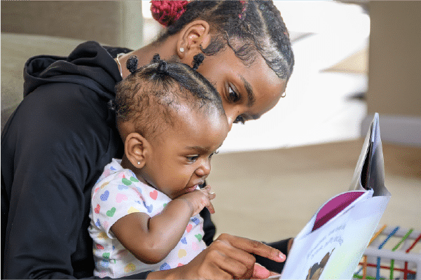 Young woman with braided hair reads a picture book to a baby with tiny pigtails who chews her fingers on her lap.