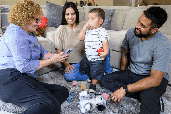 Woman showing a phone to a toddler standing on a living-room carpet while two adults sit nearby, toys scattered around.