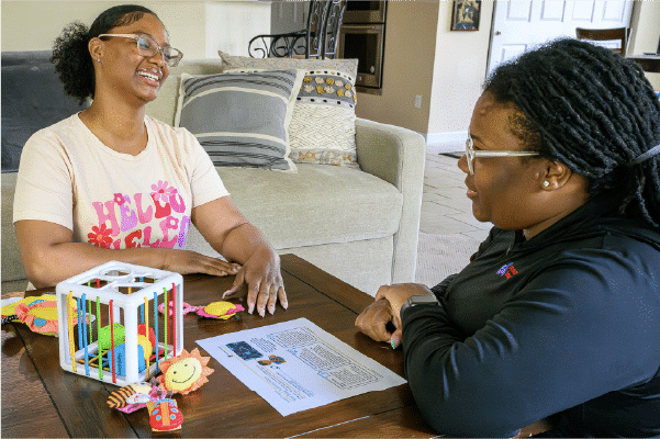 Foundational-and-Model-Implementation-Training Two women sit at a living room table smiling and talking, one wearing a light t-shirt and the other in a dark top; colorful baby toys and a printed handout lie on the table.