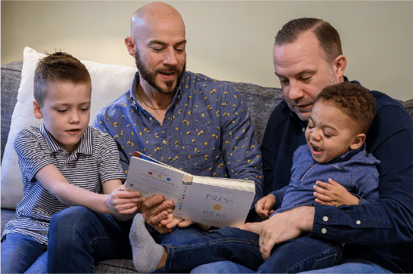 Two men sit on a couch reading a children's book aloud to two young boys, one boy on each man's lap, all smiling and engaged.