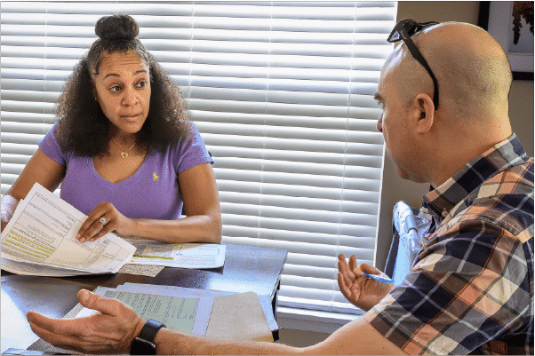 Heroes-At-Home Two adults sit at a table covered with papers and forms, the woman in a purple shirt holding documents and explaining something to the man in a plaid shirt who gestures with a pen.