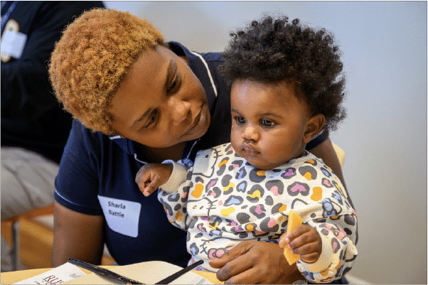 Interactions-Across-Abilities Woman with short blonde hair holding a toddler wearing a heart-patterned sweater and holding a cracker, seated at a table.
