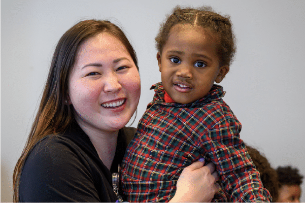 Smiling woman holding a young child wearing a red and green plaid shirt against a neutral background