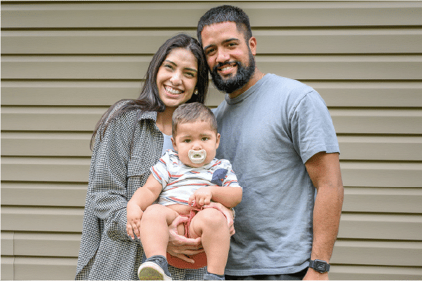 Partnering-With-Parents-Through-Screening Smiling young couple holding a baby with a pacifier in front of beige siding, mother on left and father on right.