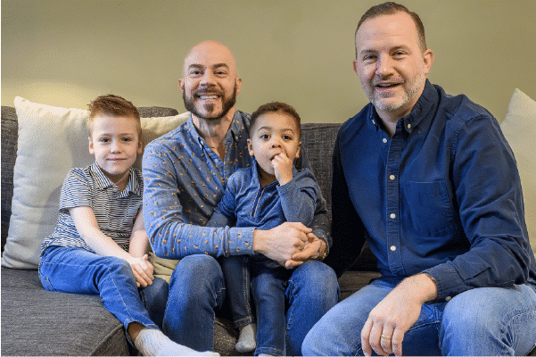 Two men and two young boys sitting closely on a couch, all smiling except the toddler who is sucking a finger, casual clothing and neutral living-room background.