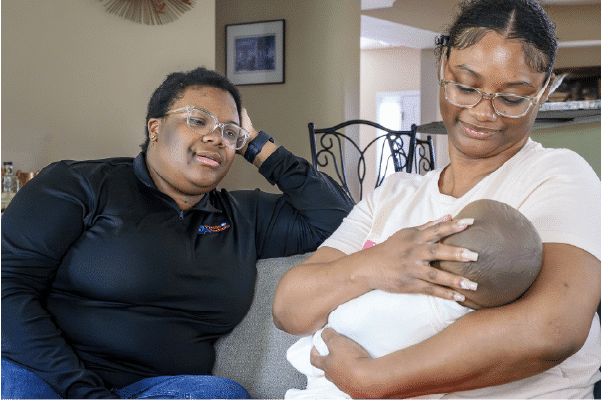 Supporting-Care-Providers-Through-Personal-Visits Two women sitting on a couch in a living room, one cradling and looking down at a swaddled baby while the other watches with a relaxed smile.