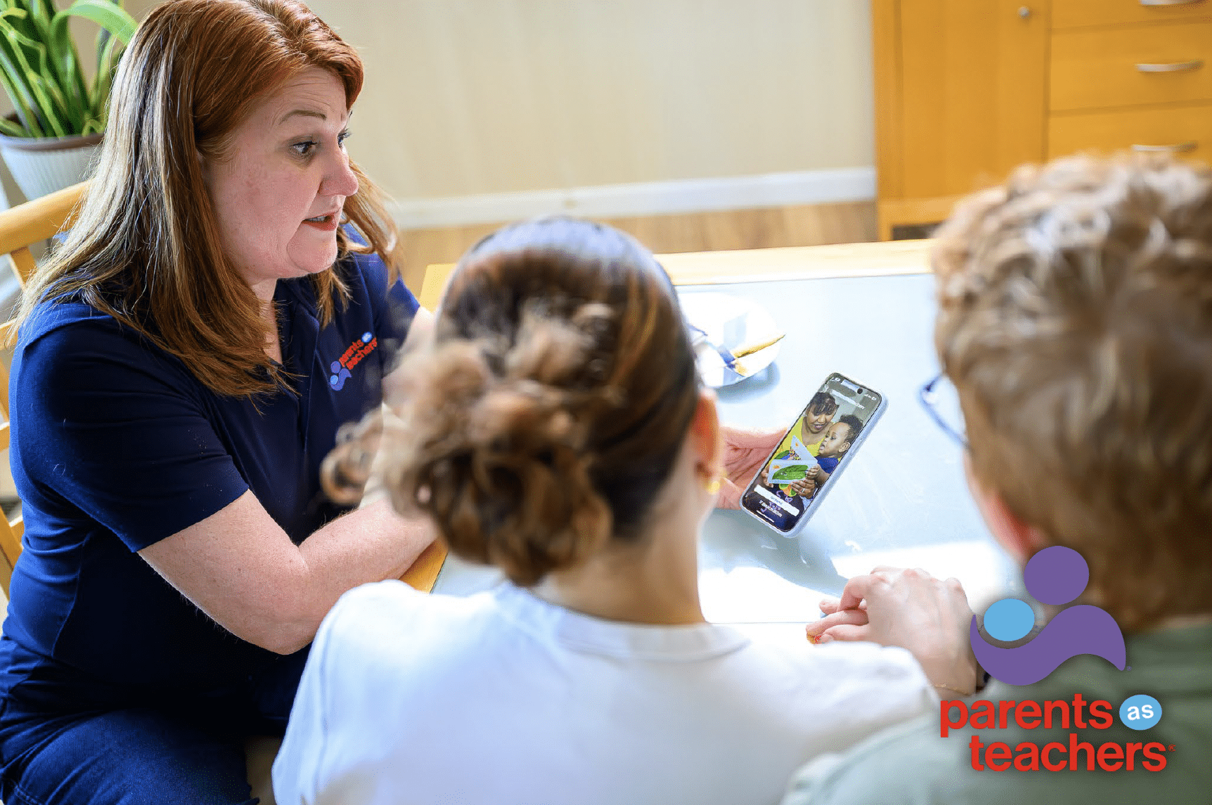 Woman in a navy polo showing two seated people a parenting app video on a smartphone at a kitchen table, Parents as Teachers logo visible.