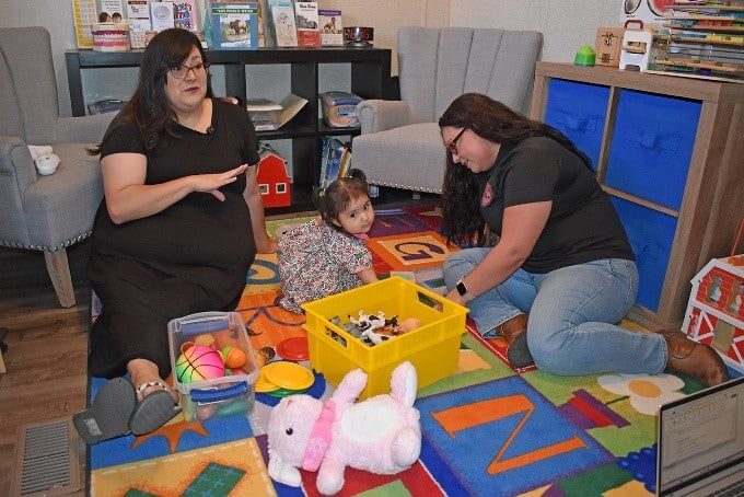 Two women and a young child sit on a colorful play mat with toy bins and plush animals in a classroom-like play area