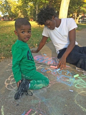 Young child in green clothes looking at camera while an older person in a white T-shirt draws colorful chalk art on a sidewalk in a park.