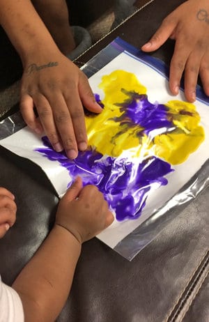 Adult and child hands finger-painting yellow and purple paint smeared on a white sheet inside a plastic bag on a dark surface.