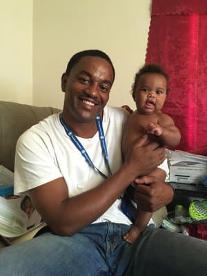 Smiling man in a white t-shirt sitting on a couch holding a laughing baby wearing no clothes, indoor living room setting with red curtain.